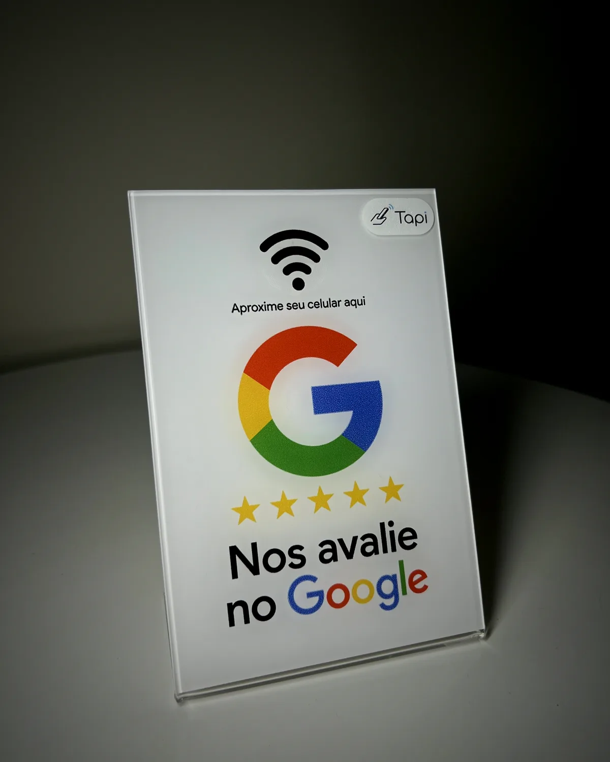 Plaquinha de acrílico branco 'Nos avalie no Google' com as cinco estrelas, sobre uma mesa